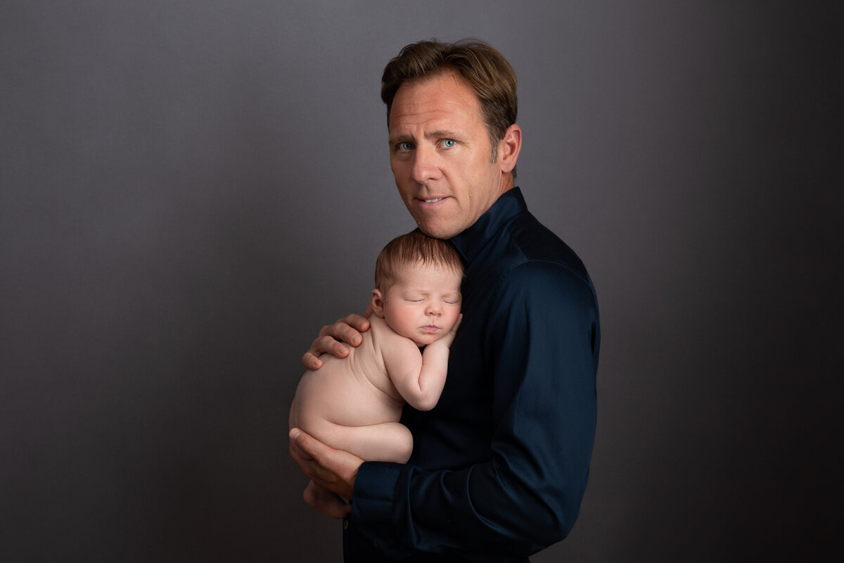 Father wearing a dark blue shirt holding his newborn curled up on his chest on a grey background.