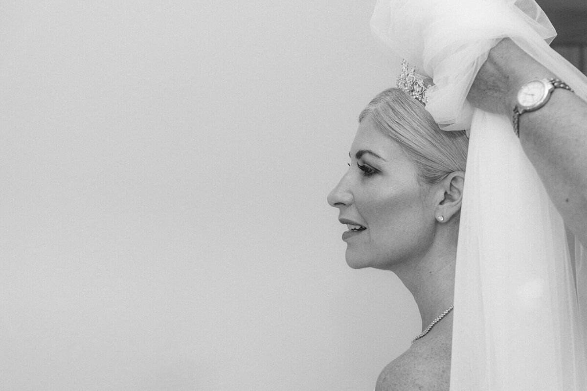 a bride, sat down and slightly off centre of the image prepares for her wedding day as her veil is fitted.