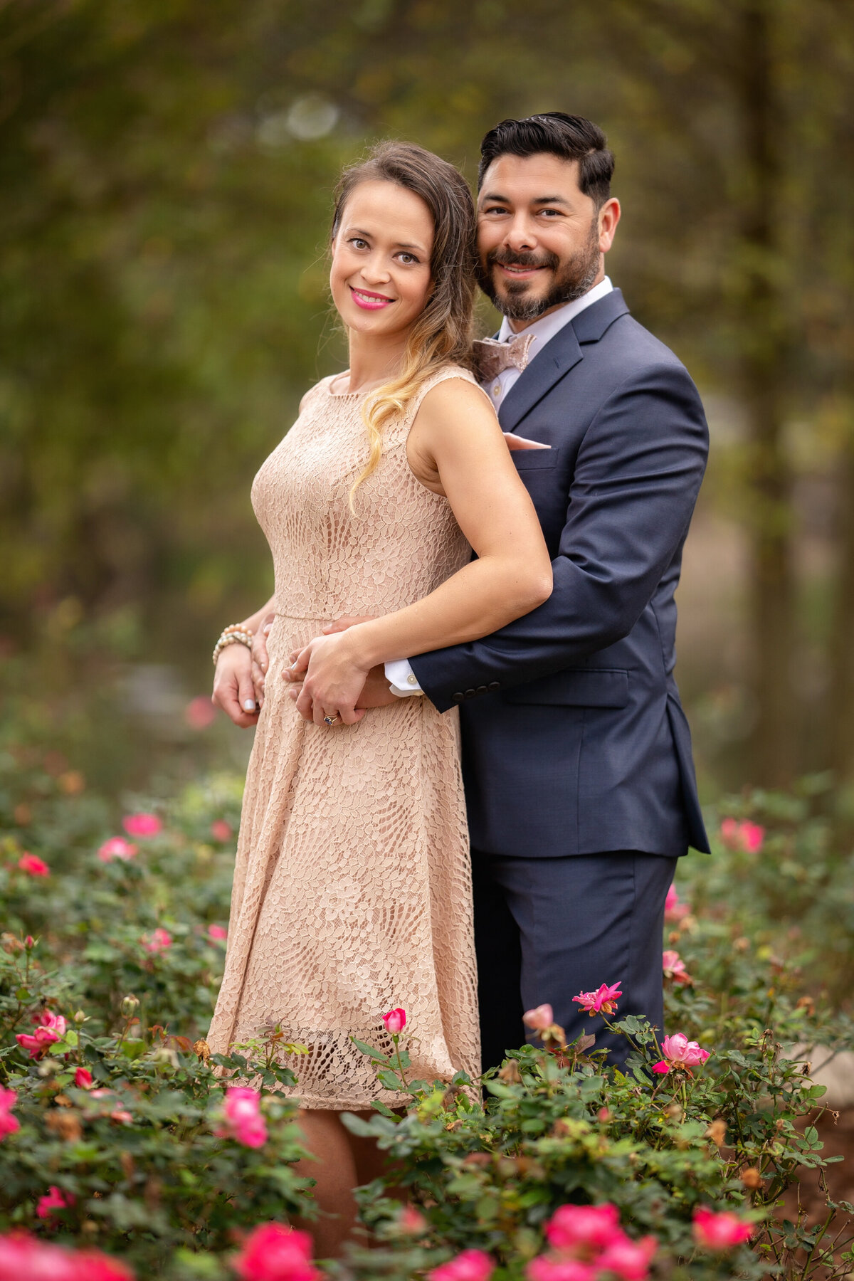 Man and woman standing in pink rose bushes.  Both are Hispanic.  He is wearing a navy blue suit and has a beard and moustache.  She has long brown hair with blonde ends and is wearing a blush dress.  She is leaning with her back to him and they are holding hands.  There are blurry trees in the background.  They are smiling at the camera.