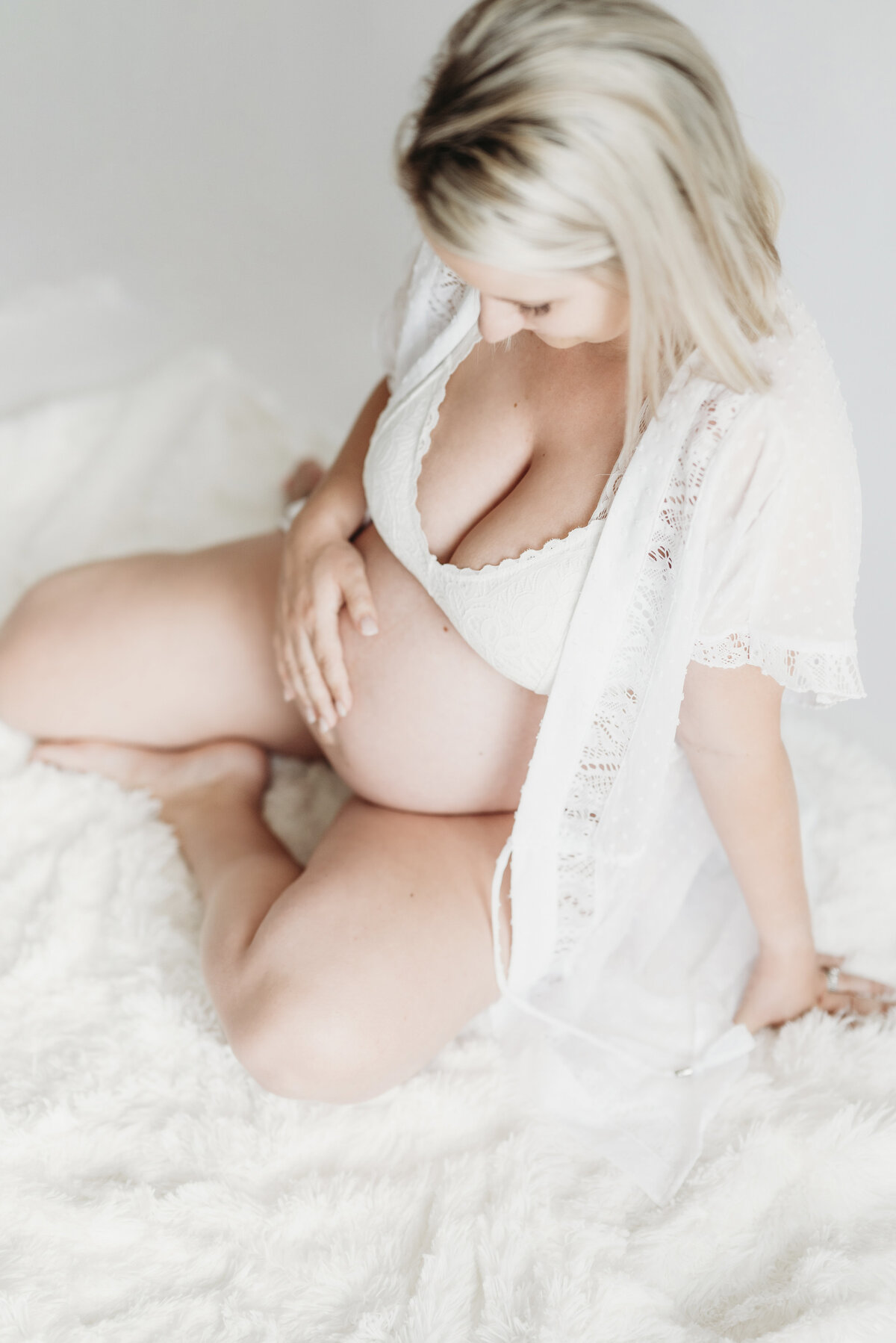 Maternity boudoir session featuring an expecting mother seated on a bed in a white lace robe.