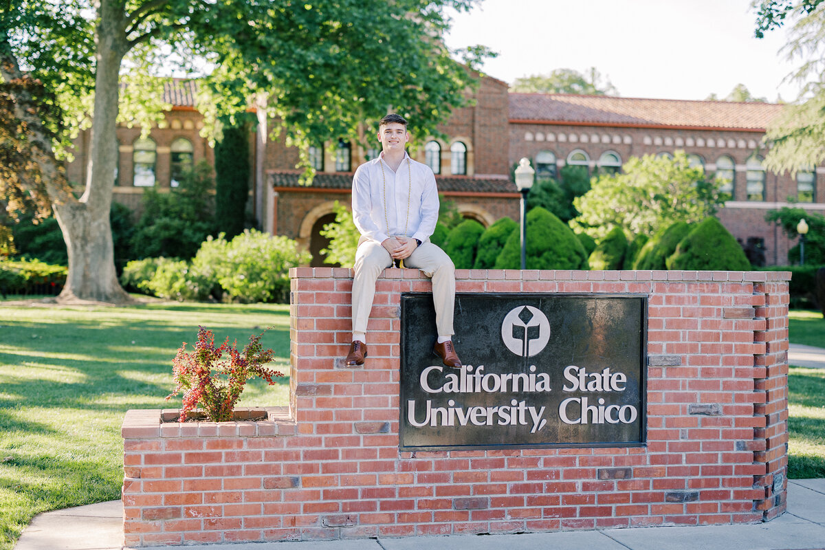Chico State senior graduate wearing cap and gown, celebrating graduation on campus with Chico State University buildings and greenery in the background.