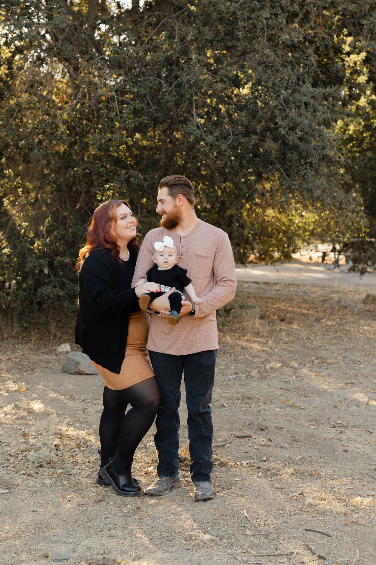Family standing together outdoors with baby in mother’s arms, surrounded by tall trees. Photographed by Yucaipa family photographer Kaitlyn Dawn Photography.