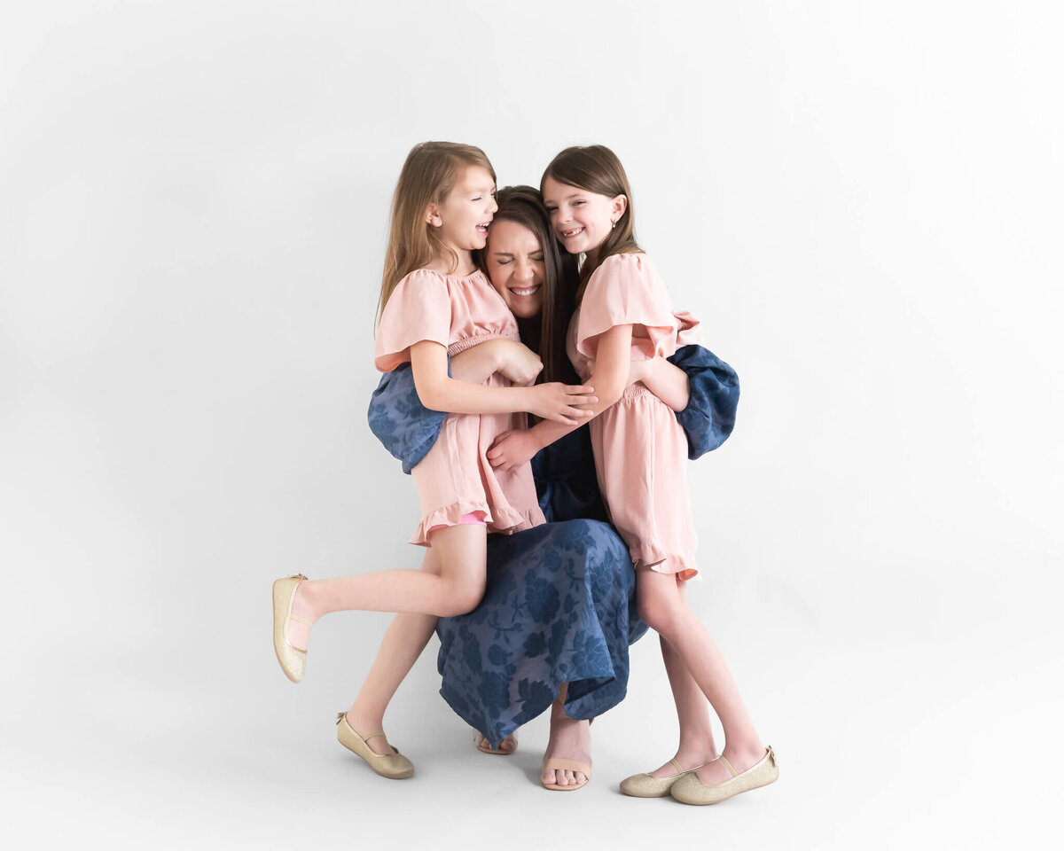 mom  on ble dress crouching down hugging her 2 daughters both wearing matching pink drresses