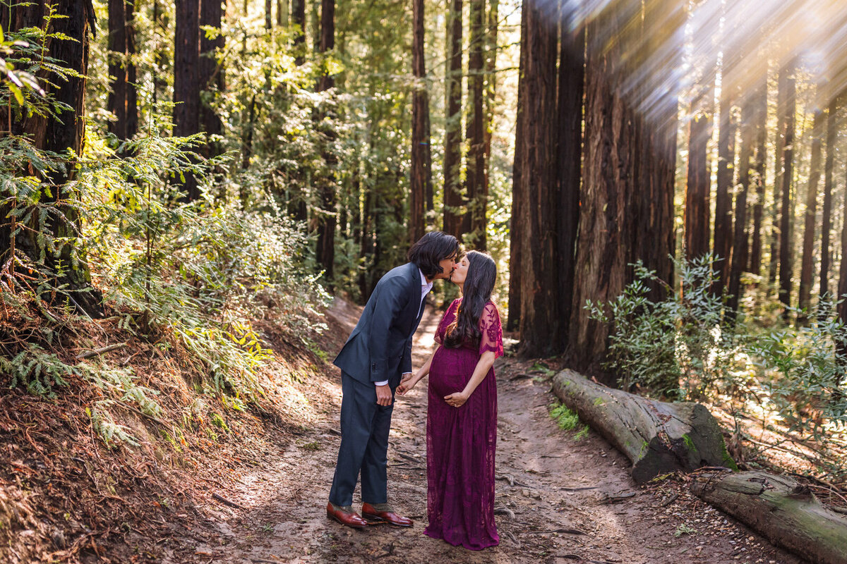 pregnant woman in purple dress standing in Oakland redwoods as husband leans in to give her a kiss