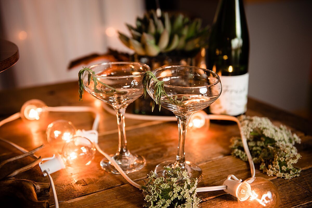 Two gold rimmed coupe glasses sit on a wine barrel surrounded by a strand of lights with greenery, a succulent and a green wine bottle.