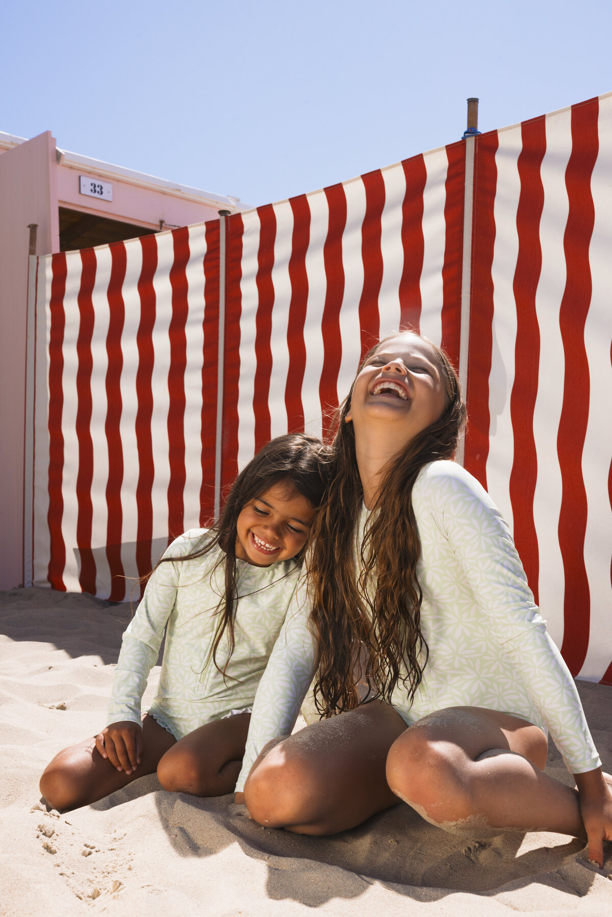 Two girls sitting and laughing in the sand in front of red and white striped beach windbreakers, both wearing matching mint green Swim Essentials UV swimwear with a leaf print.