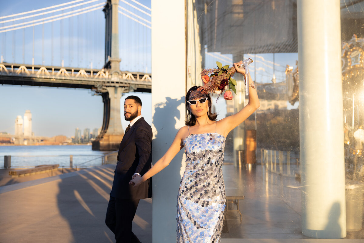 The couple poses together at the base of the Brooklyn Bridge, surrounded by urban textures and history.