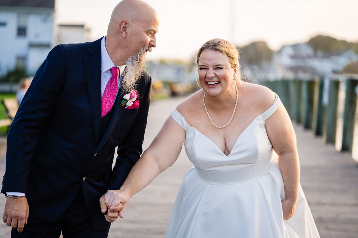 The bride laughs joyously while holding hands with the groom on a waterfront boardwalk