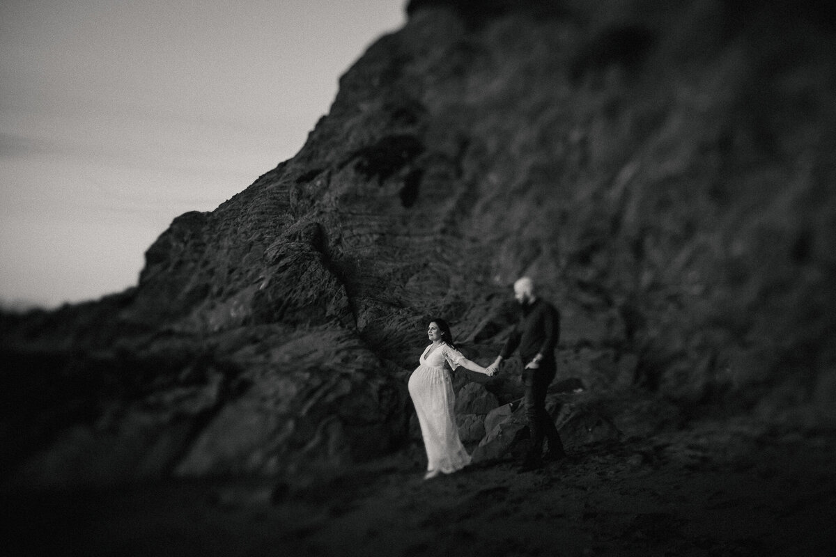 San Francisco maternity couples photography in black and white.  Pregnant mom in white dress leads dad walking on rocky coast