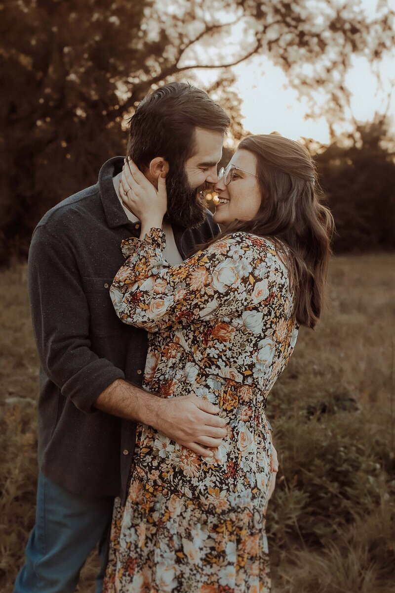 A couple embraces outdoors with the woman touching the man's beard. They stand in a grassy area with trees in the background.