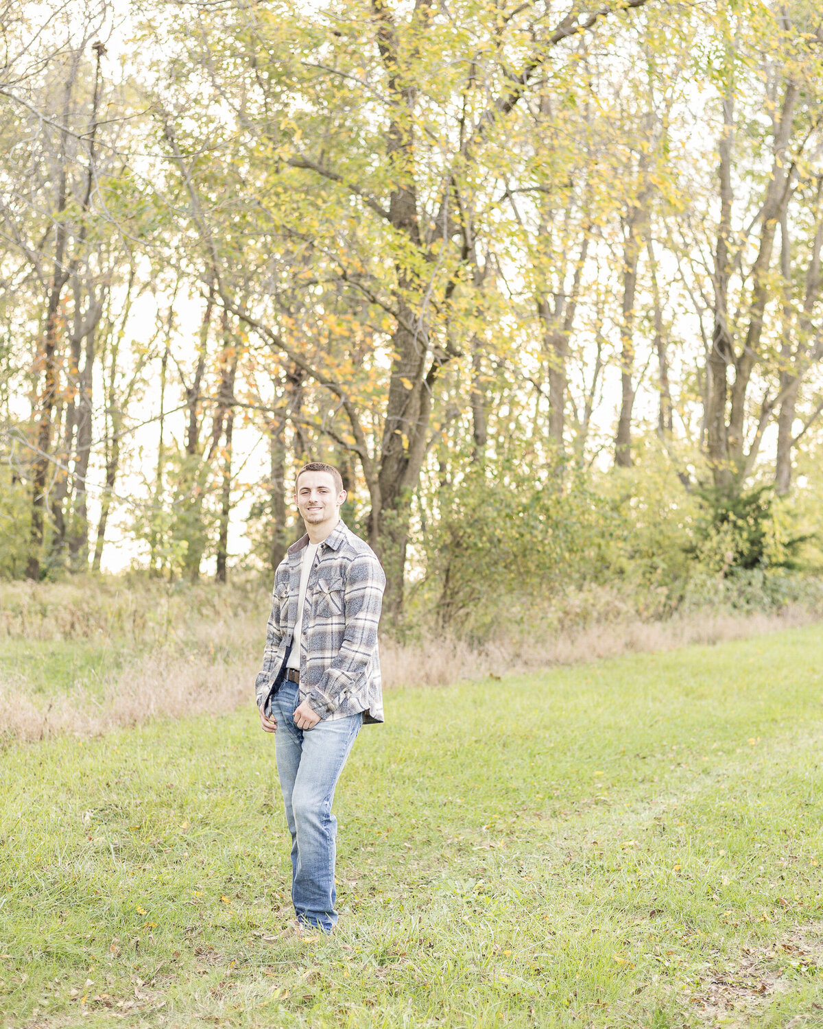 high school senior male, standing in grass , with trees behind him, wearing blue jeans, flannel jacket, light tan shirt, looking at the camera smiling.