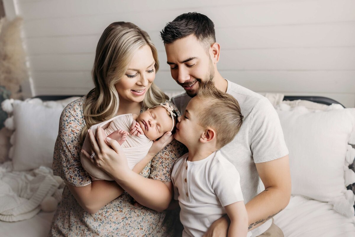 Parents posing with newborn baby as toddler brother kisses babies head in a white bedroom studio setting