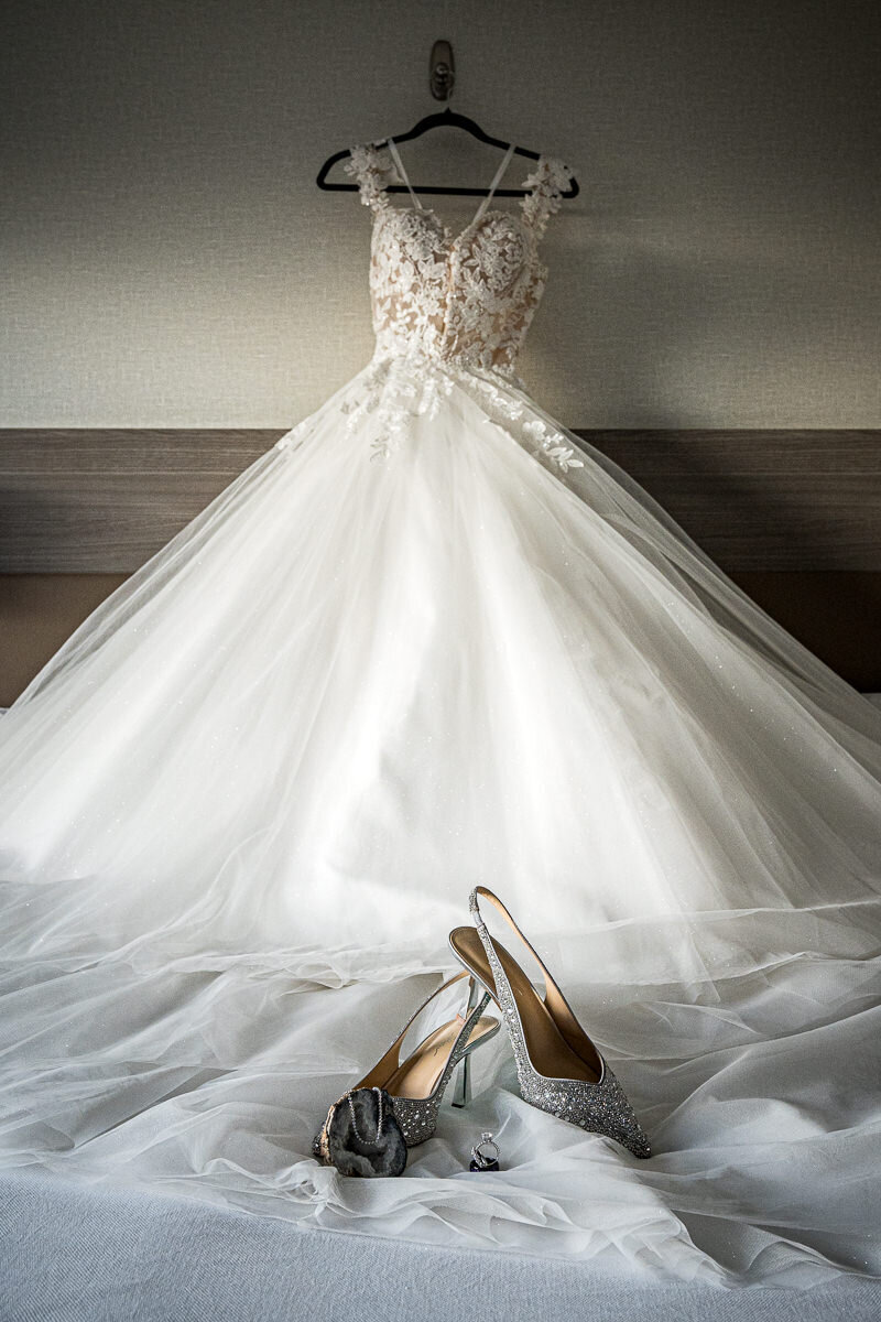 A beautifully styled shot of the bride's dress hanging gracefully, with her shoes and wedding rings elegantly displayed in front, captured at the AC Hotel Pleasanton to highlight the romantic details of the wedding day.