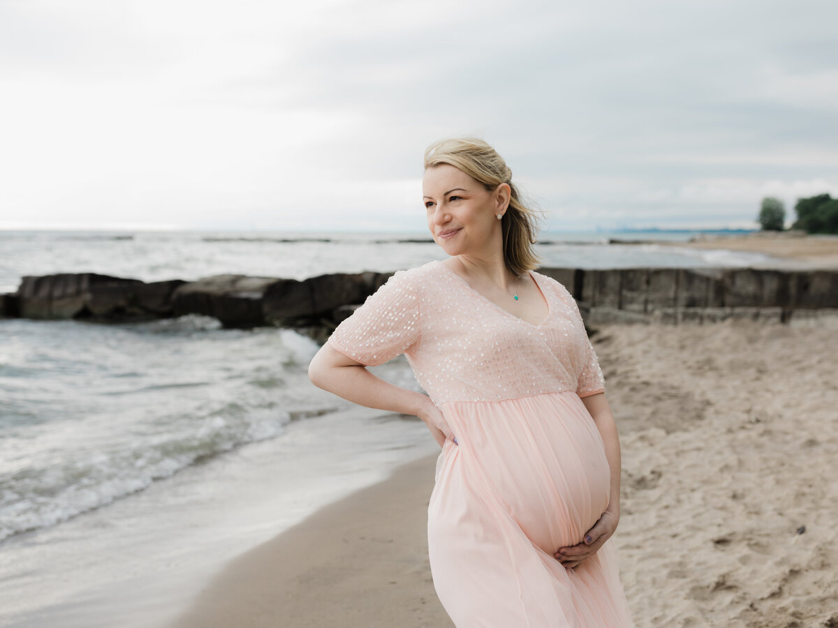 pregnant mother in pink maternity gown holding baby bump on beach for maternity photoshoot cleveland maternity photographer