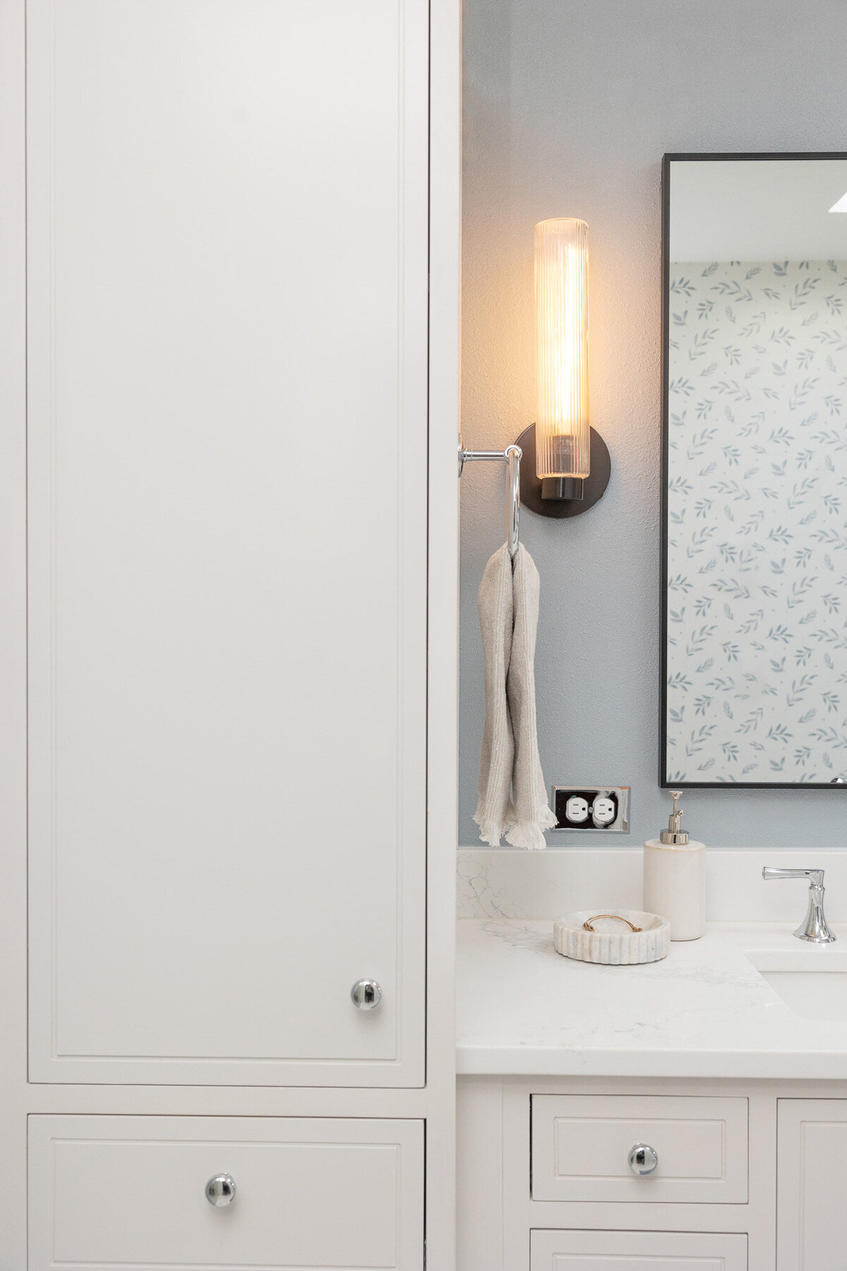 White linen cabinet, white vanity with silver pulls, black mirror and black illuminated sconce, white countertops, blue walls