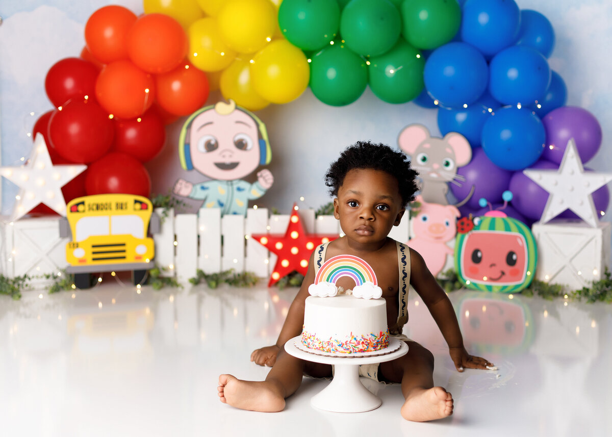 Cocomelon themed cake smash in West Palm Beach, FL and Jupiter, FL. newborn and cake smash photographer. Black baby is sitting behind a sprinkle cake looking at the camera. In the background, there is a rainbow ballooon arch and cutouts of Cocomelon characters.