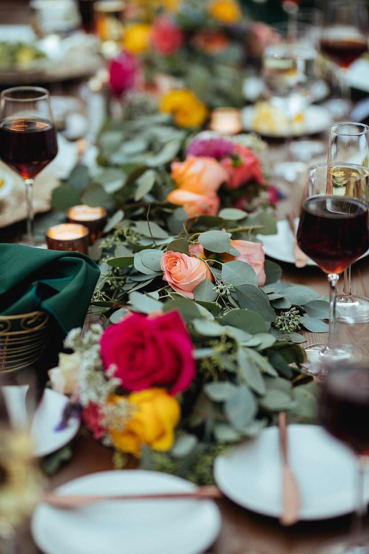 long farm table set with bread and butter plates with small gold butter knives and a long garland of greenery accented by hot pink, yellow, orange and peach roses, low candles in gold votive holders and stemmed wine glasses with red wine
