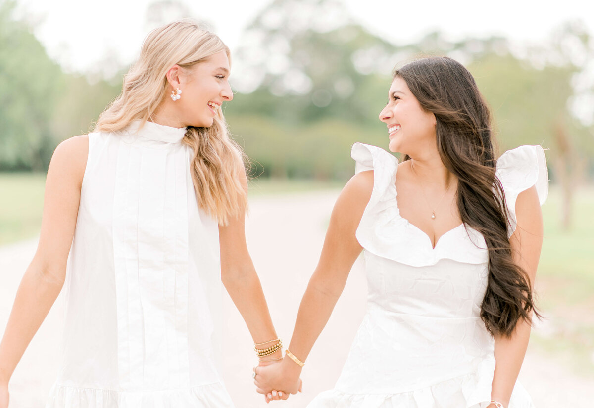 High school senior girl friends hold hands and walk along a gravel path, smiling at each other.