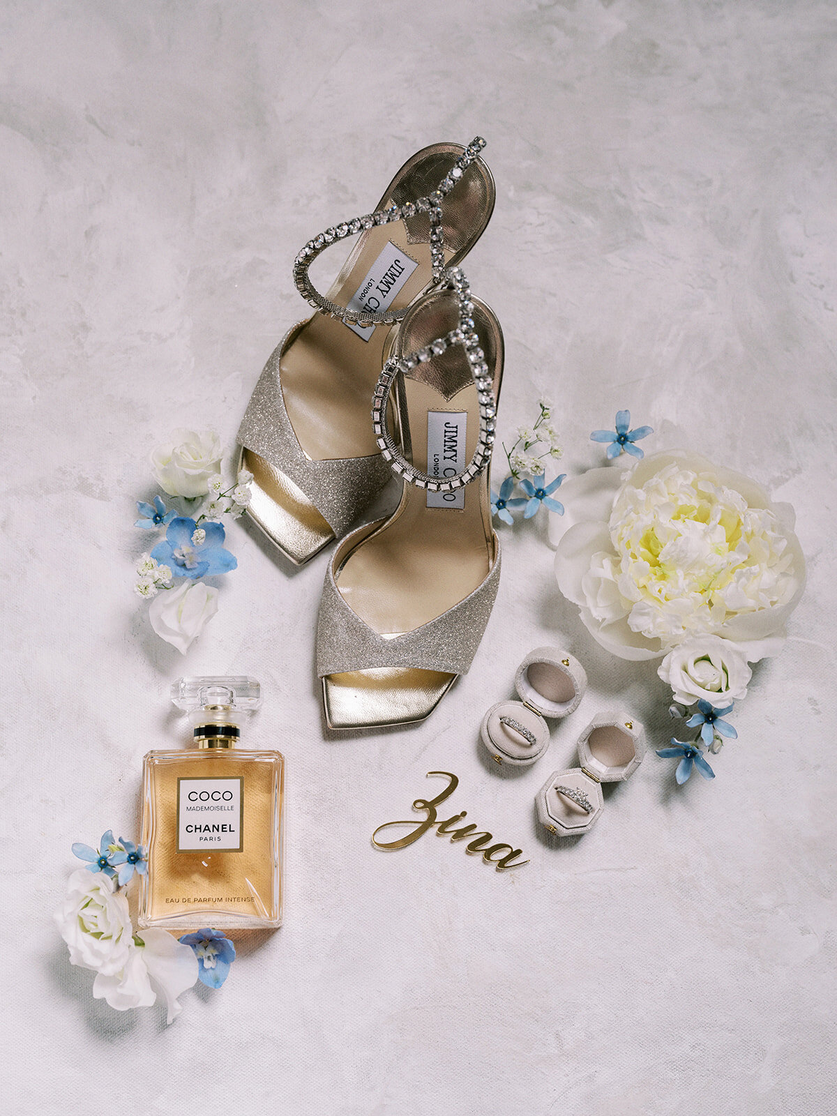 A pair of glittery high-heeled shoes with ankle straps, a bottle of Chanel perfume, white flowers, small blue flowers, and a “3ina” label arranged on a light textured background, capturing the elegance and charm perfect for a classic Calgary wedding.