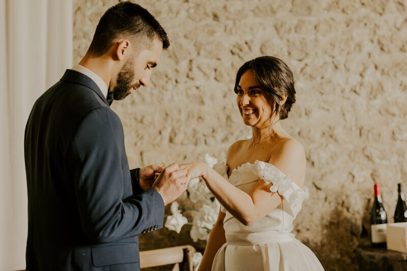 Mariée passant l'alliance au doigt de la mariée, posant pour le shooting photo. Voilage et mur en pierre en arrière plan.