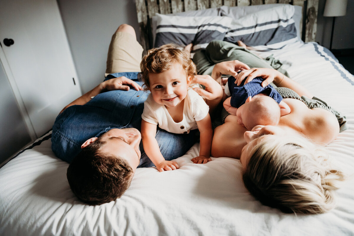 Newborn Photographer, a young family of 4 lays on the bed, an older toddler sister smiles