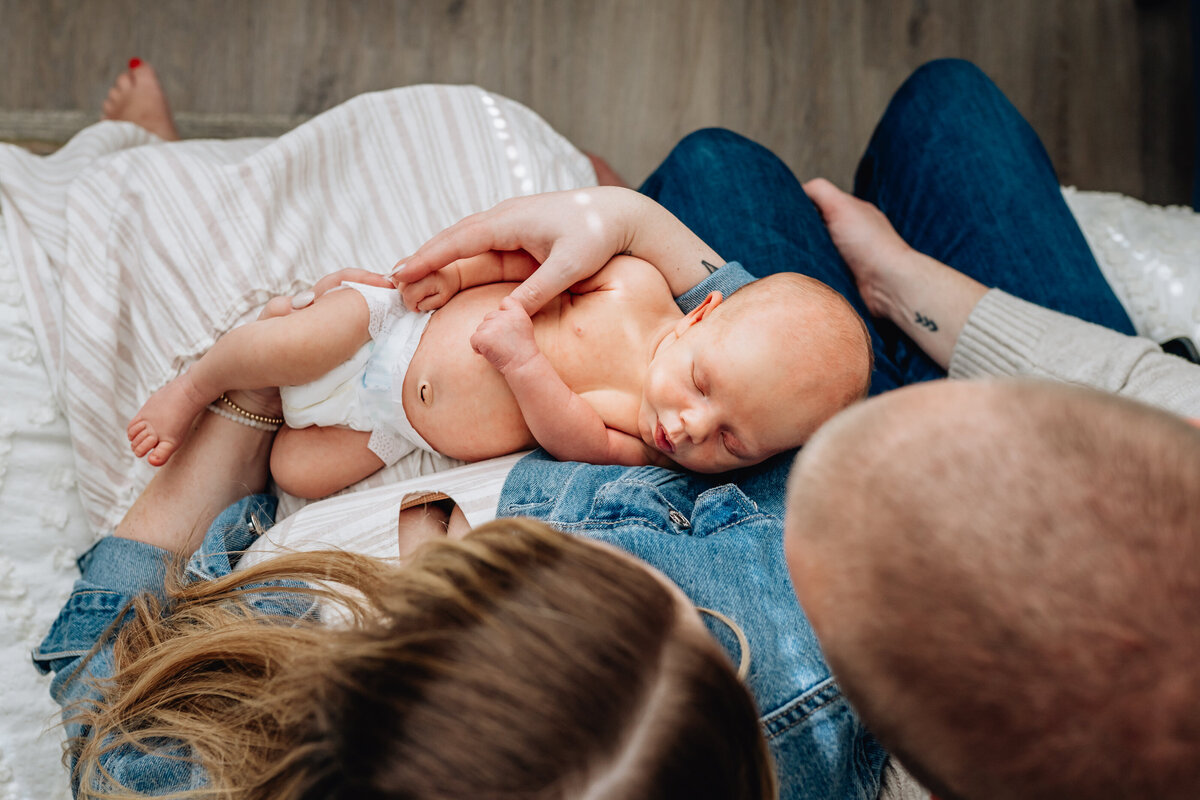 A top-down view of a newborn baby resting on their parent's lap, with one parent cradling the baby's head while the other parent supports the baby's body. The baby is wearing a diaper and appears to be asleep.