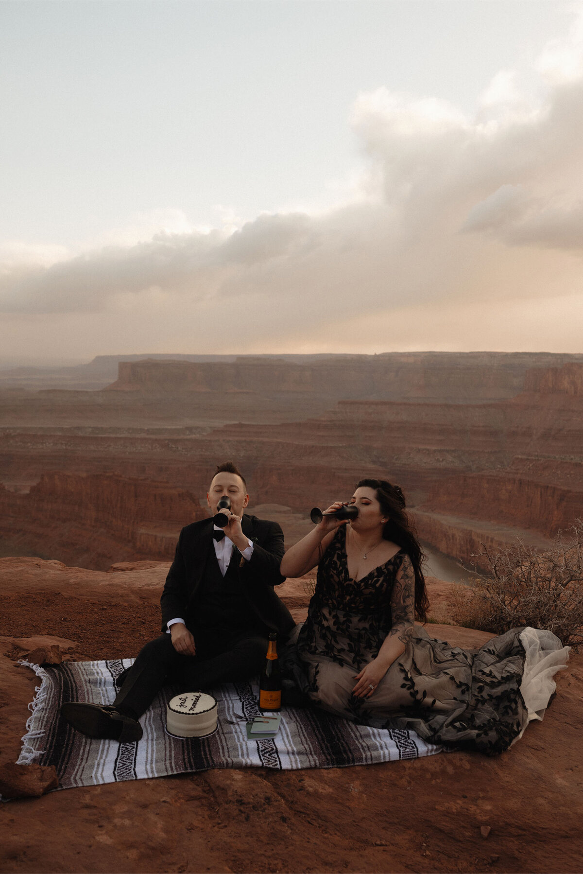 Moab, Utah elopement couple sip on champagne while sitting on a blanket and eating cake after their ceremony at Dead Horse Point State Park in Moab