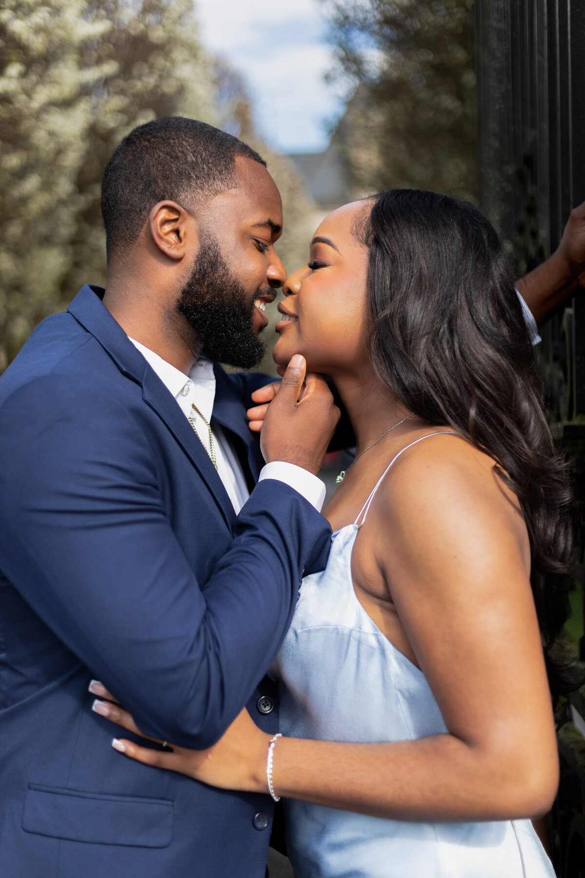 Ochre Court Mansion  | Kelsey Sheehan Photography Timeless Rhode Island Weddings | A couple stands close outdoors, smiling and touching noses. The man in a blue suit gently holds the woman's chin. They appear happy and in love.