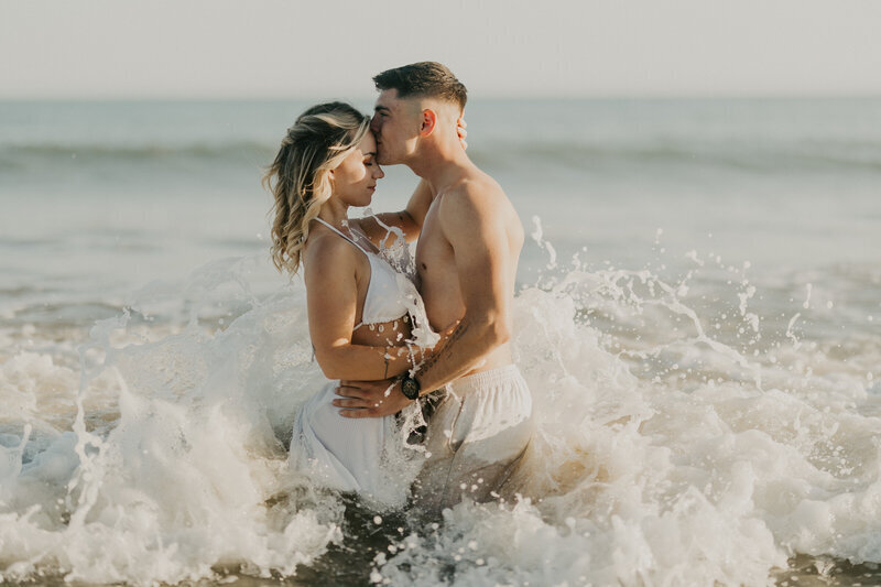 Couple enlacé dans le flot des vagues. L'homme fait un bisou sur le front de sa chéri. Capturée par Laura Termeau photographie.