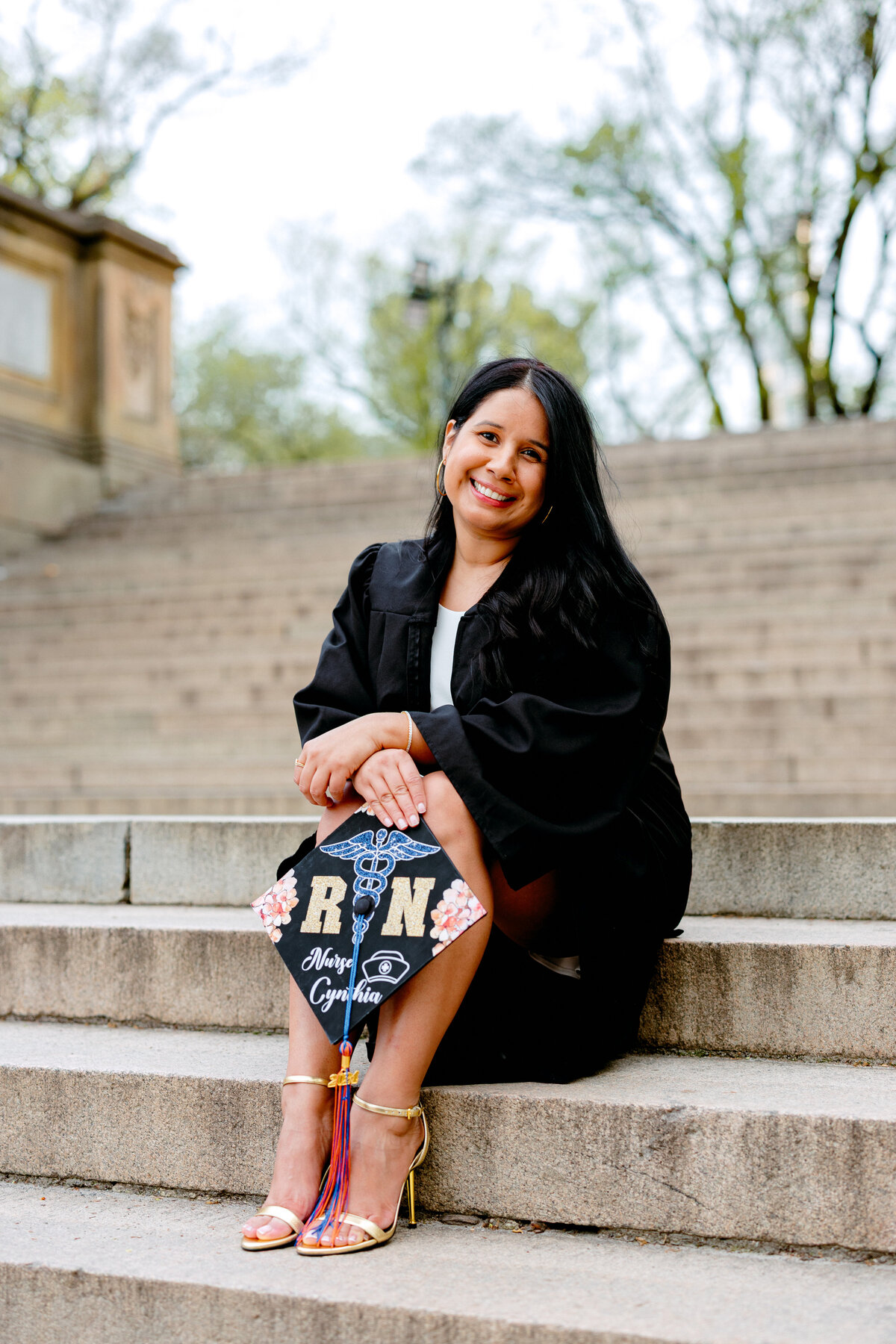 Hostos Community College graduate smiling while sitting on the steps at Central Park and holding decorated cap.