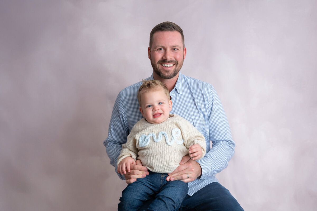 Smiling toddler wearing a ‘one’ sweater sits on the leg of a smiling man in a light blue button-up shirt.