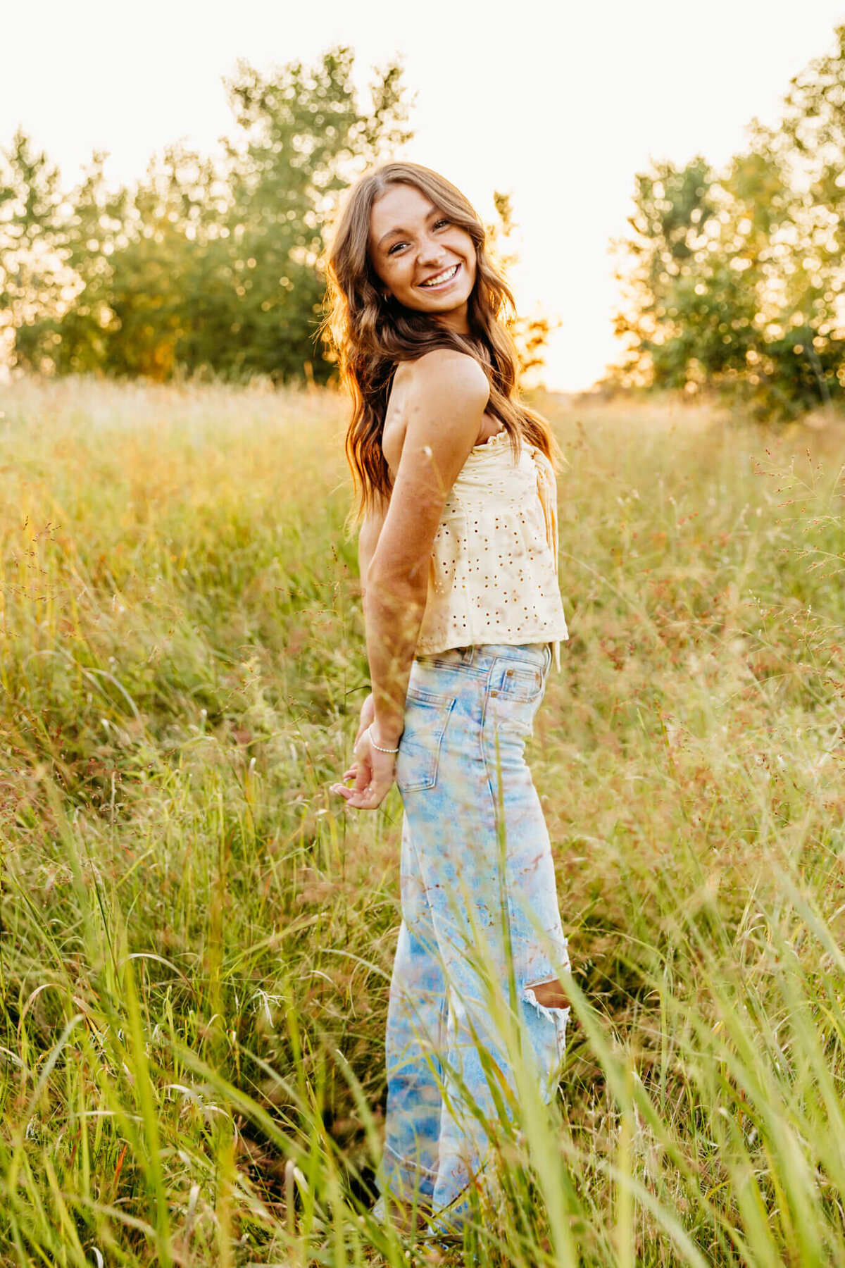 brown haired teen girl in a tank top and jeans standing with her hands behind her back and laughing in a field near Appleton Wi