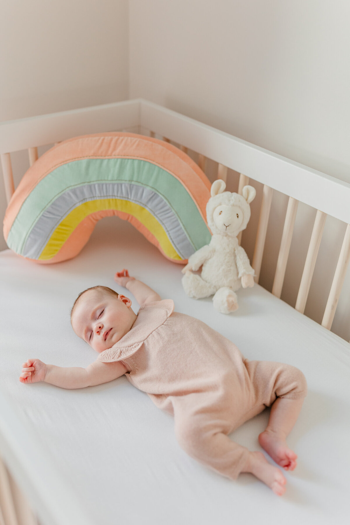 Boston newborn baby in soft pink outfit sleeping with her hands up by her head in a crib decorated with a rainbow and llama plushie