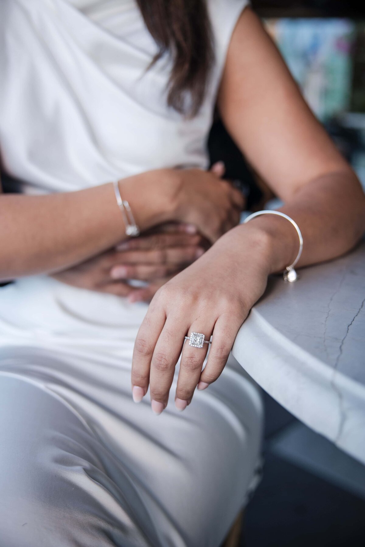 A close-up photo of an elegant bride-to-be’s hand resting gently on a marble table, showcasing a stunning emerald-cut diamond engagement ring. She wears a modern white dress with minimal silver jewelry, while her partner lovingly embraces her from behind. This intimate moment was captured during a hotel rooftop bar engagement session in St. Augustine, Florida. The image highlights the refined, classy aesthetic of the couple and the timeless style of the session—photographed by a St. Augustine engagement photographer known for candid, true-to-color imagery.