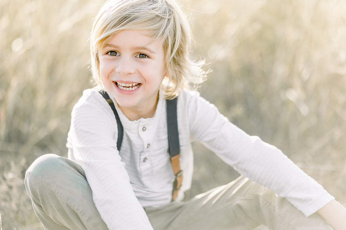 Close up photo of a young boy sitting down in a field smiling.