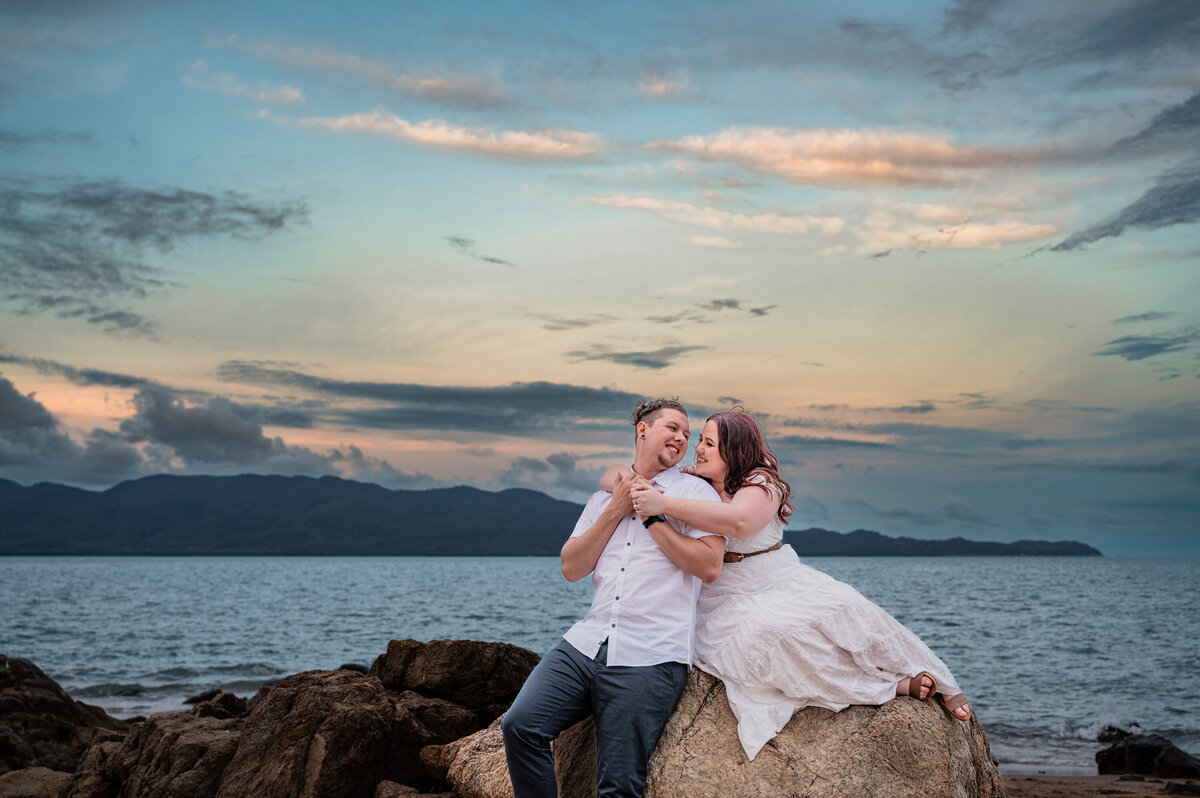 man an woman sitting on a rock on the beach at sunset - Townsville Engagement Photography by Jamie Simmons