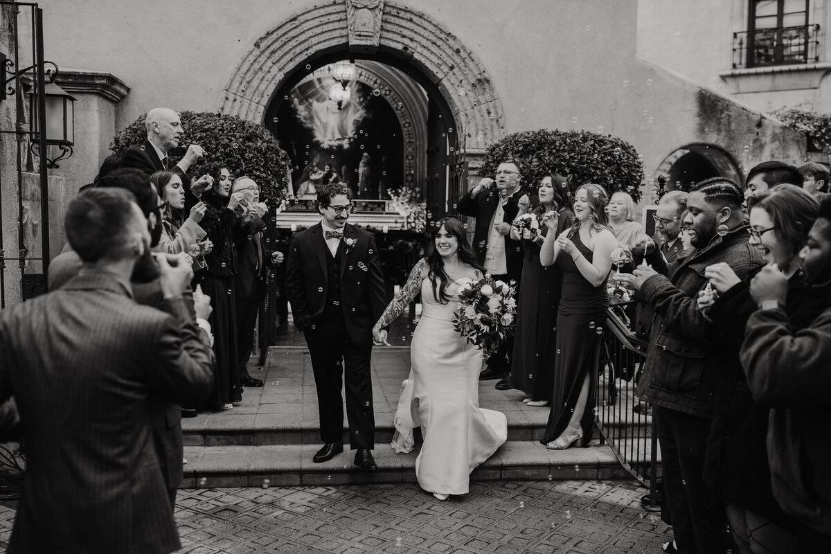 black and white photo of a couple holding hands while leaving the tlaquepaque wedding chapel while their friends and family blow bubbles for them to run through