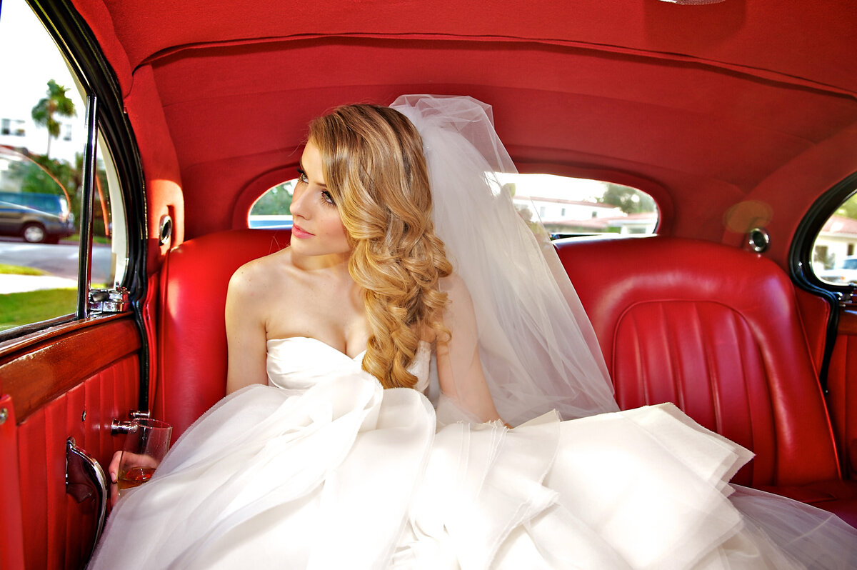 Bride sitting inside a classic car with red interior, gazing out the window.