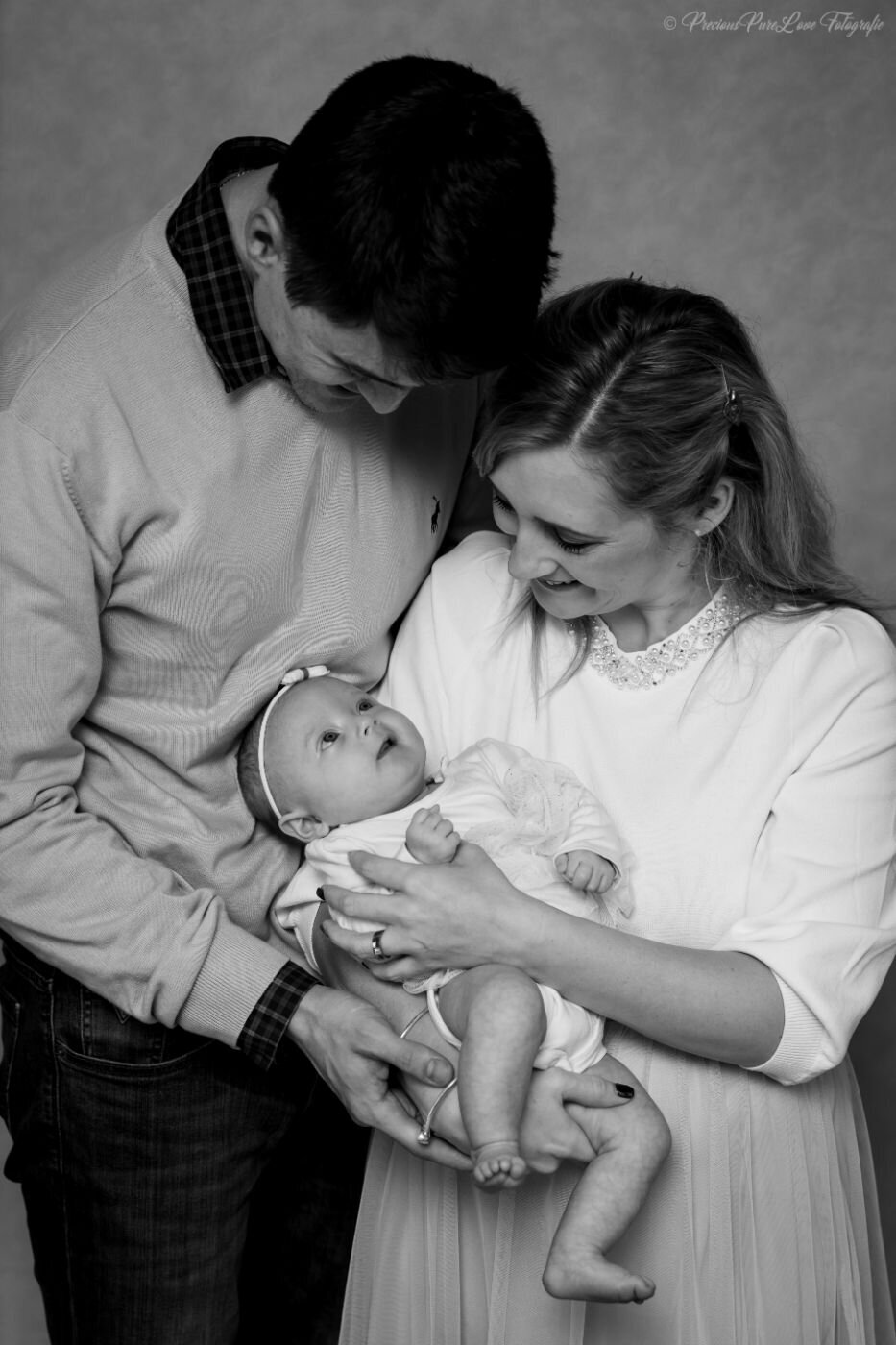  A black and white, posed family portrait of a mother, father, and their newborn baby. The mother, with a half-up hairstyle and a dress with a beaded collar, holds the baby. The father, in a sweater and jeans, leans over to look at the baby. The baby, wearing a small white bow on its head, is looking up at the parents.