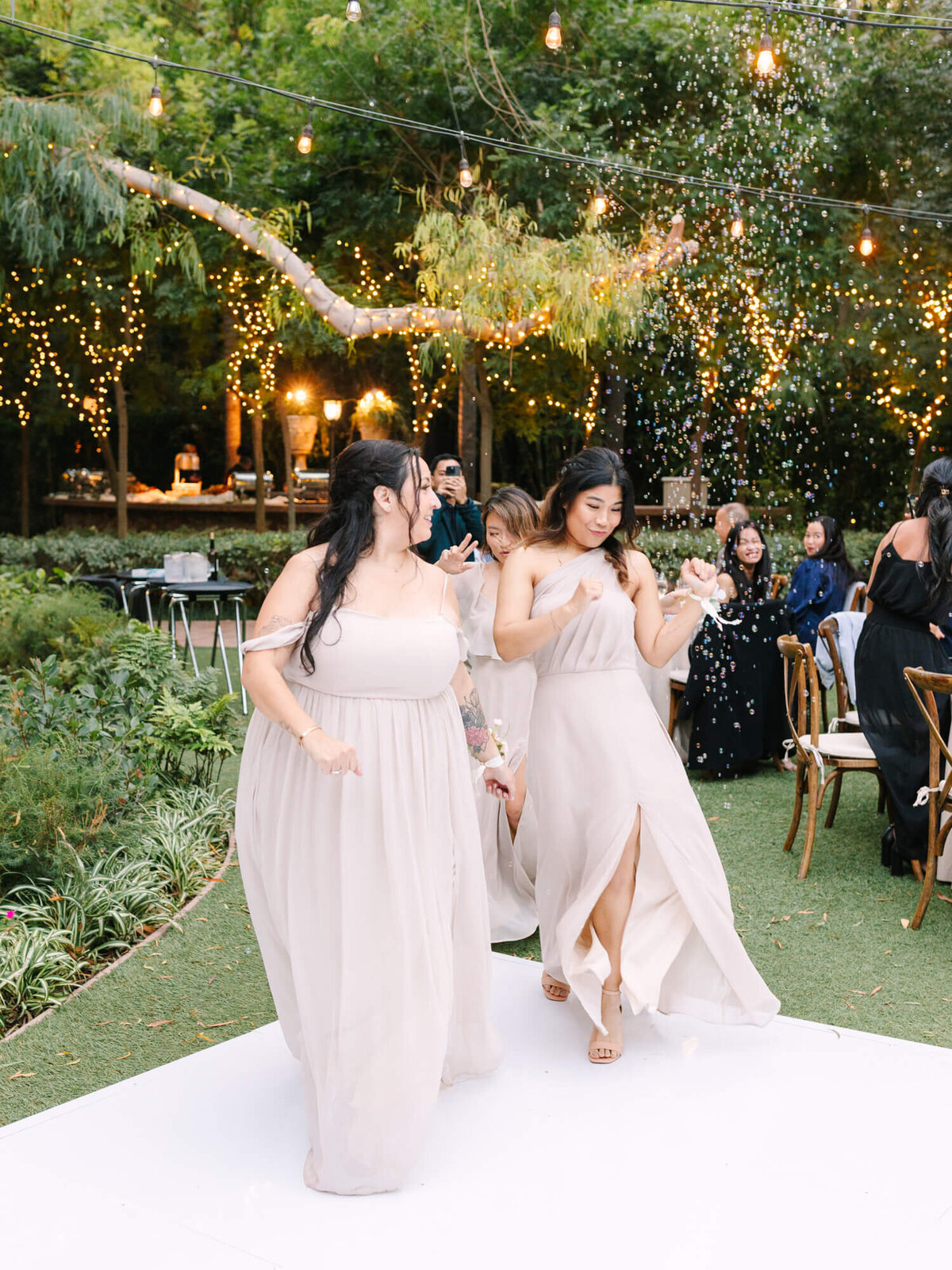 Bridesmaids in elegant pink dresses dance joyfully on a white dance floor, surrounded by greenery and softly glowing lights.