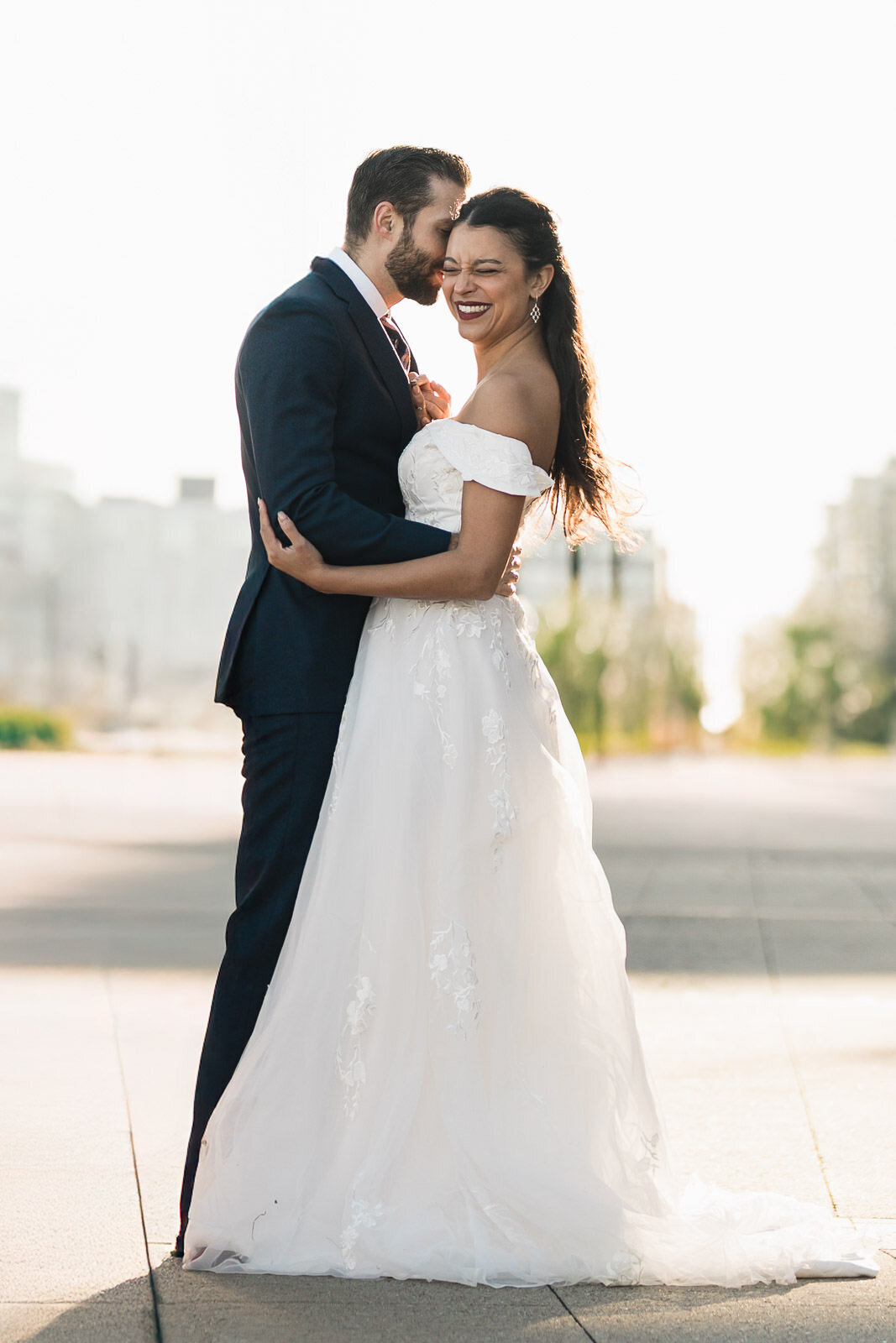 Bride laughing as the groom whispers to her during a golden-hour wedding portrait on the Jersey City waterfront, captured with soft backlighting