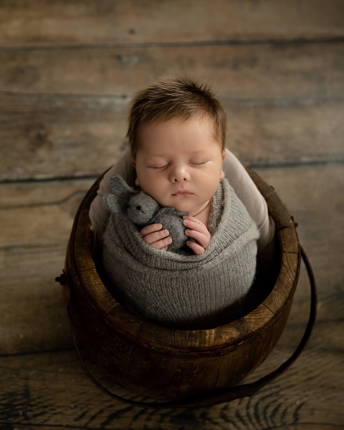 Newborn boy with lots of light brown hair sleeping in a wooden bucket holding a felt gray rabbit.