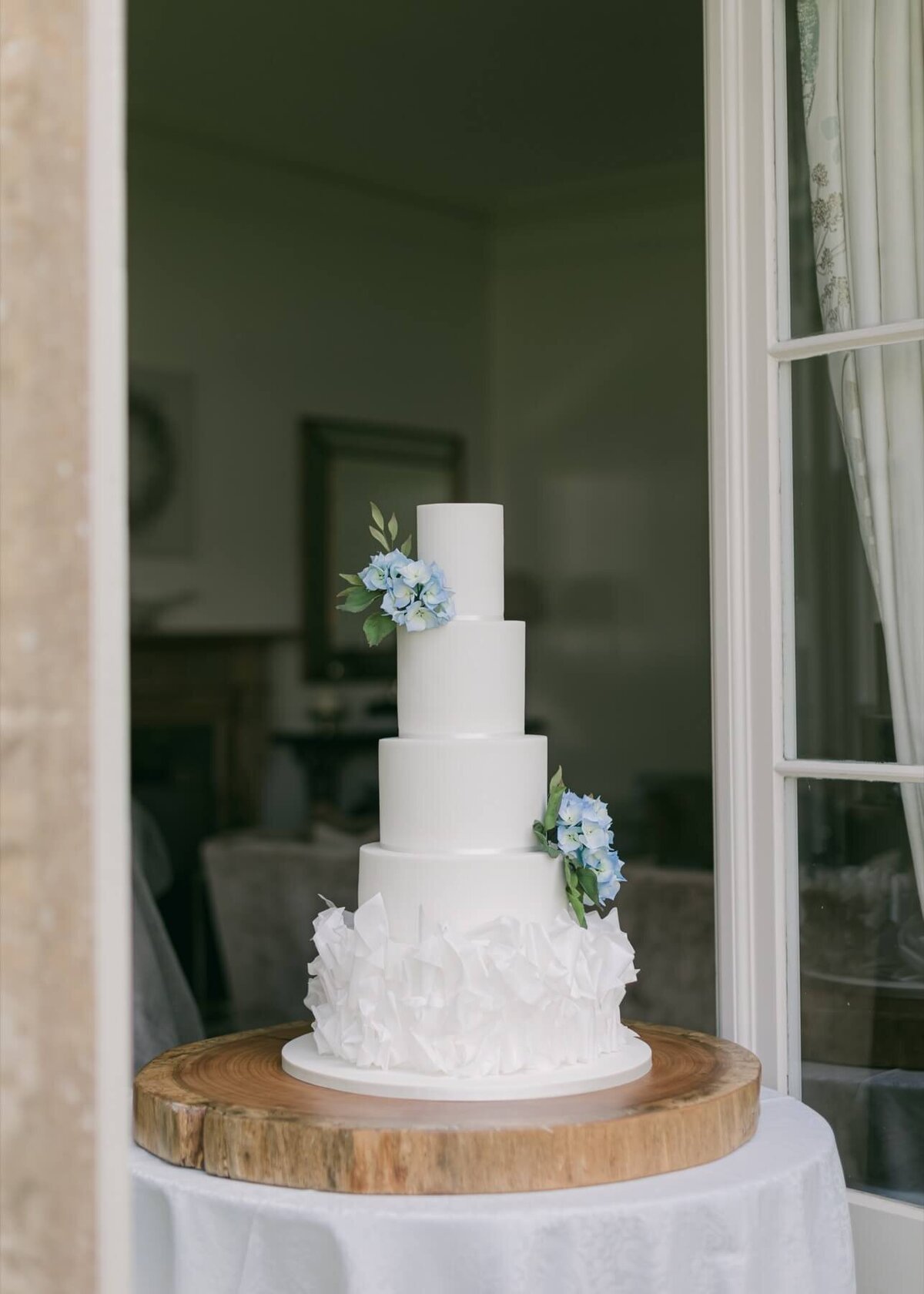 A wide shot of a white wedding cake showing ruffles at the bottom and blue hydrangea sugar flowers