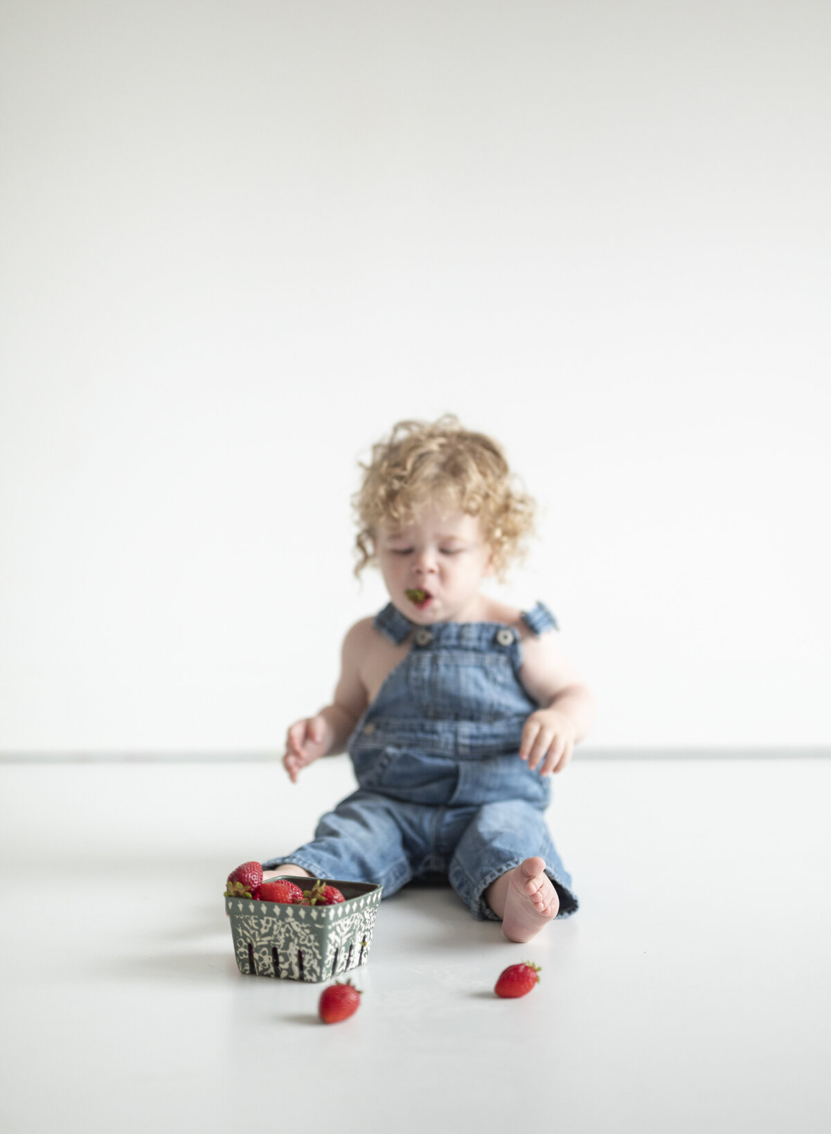 Toddler sitting alone on a white floor, focused on eating strawberries.