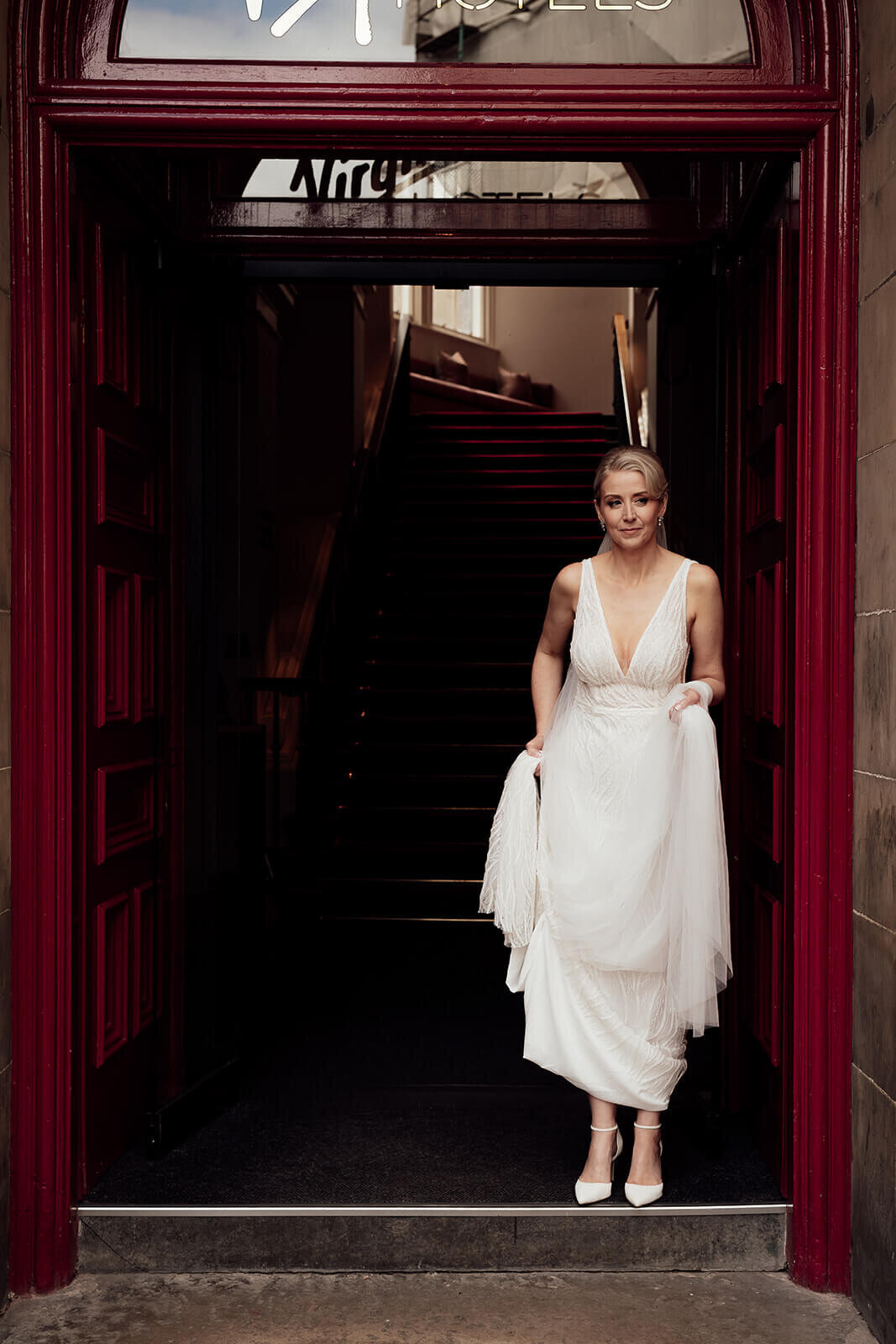 A bride stands holding her dress up in a red door way