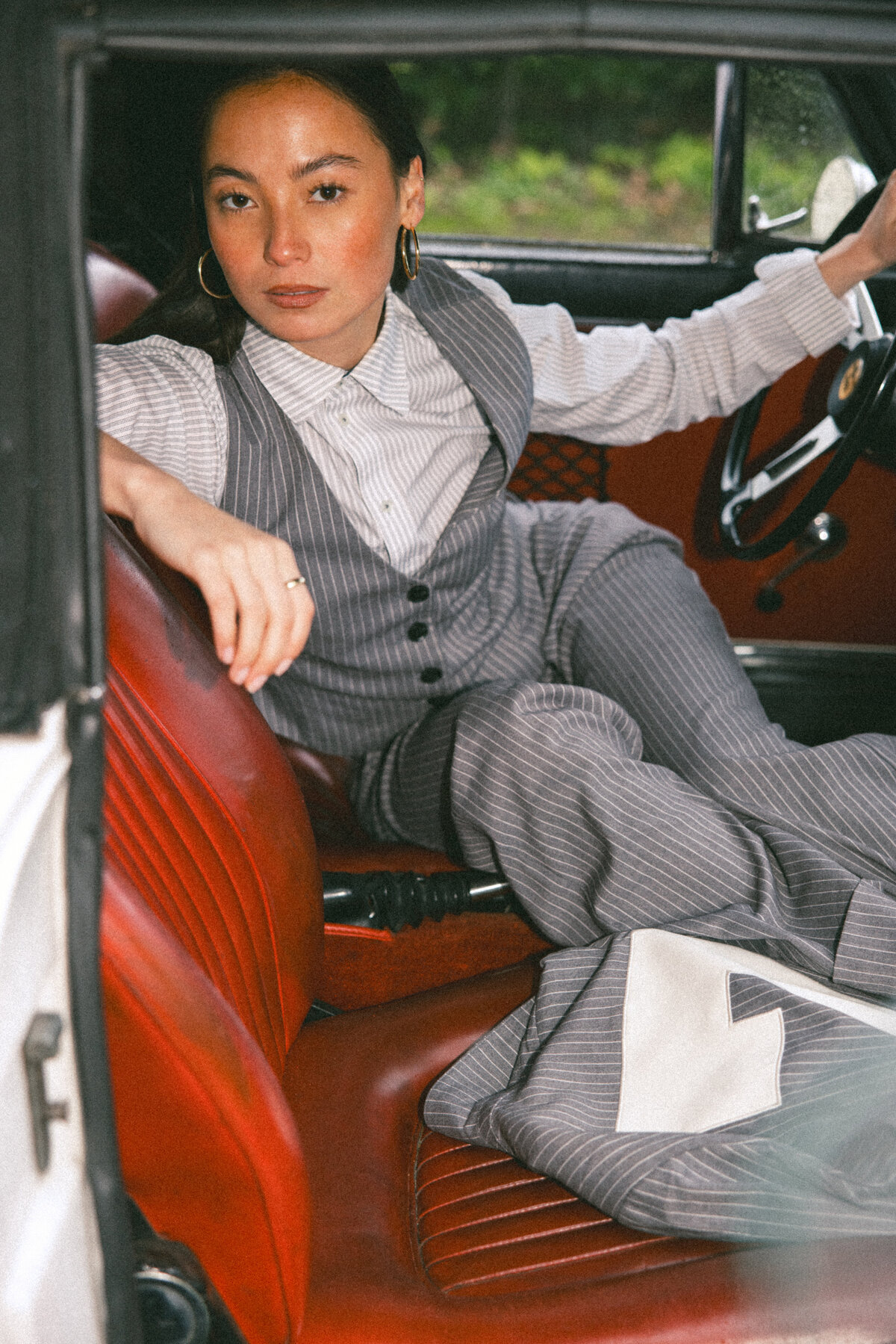A striking portrait of a confident woman seated in a vintage car, dressed in a pinstriped suit. Her assertive gaze meets the camera, embodying elegance and strength. The red leather interior of the classic car adds a rich, vintage vibe to the scene, photographed by Bo Eppink.