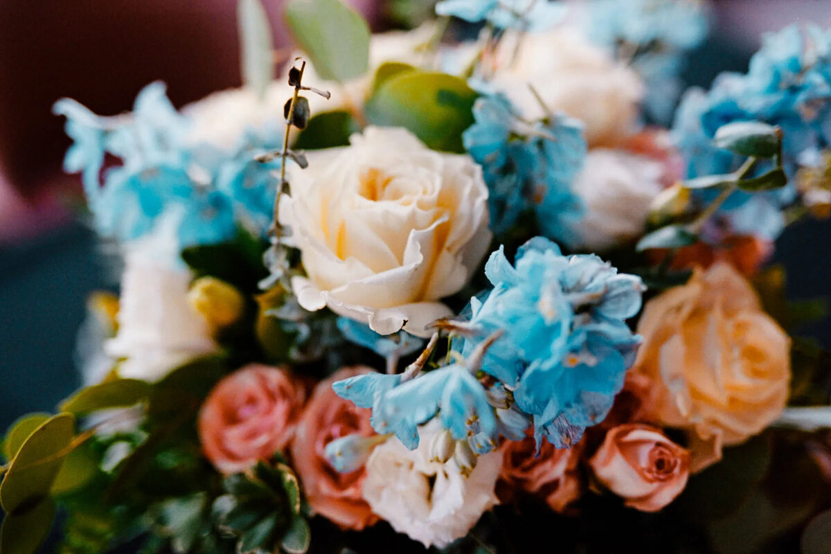 A close-up of a floral arrangement featuring cream-colored roses, peach roses, blue delphiniums, and green leaves, beautifully captured by an NJ wedding photographer with a soft, blurred background.