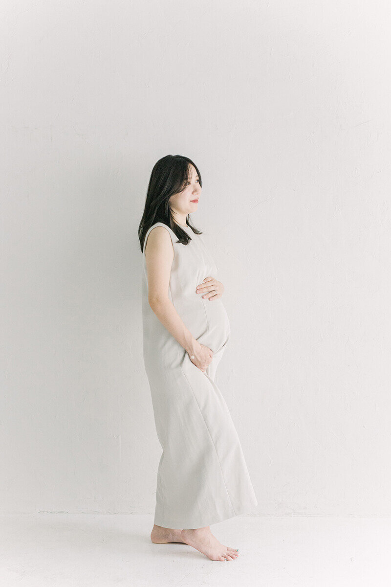 ホワイトコーデのおしゃれで美しいマタニティフォト。妊婦さんがおなかに手を当てて、外を眺めている素敵な写真。新宿のスタジオにて、Yukika Ishikawa Photographyが撮影。