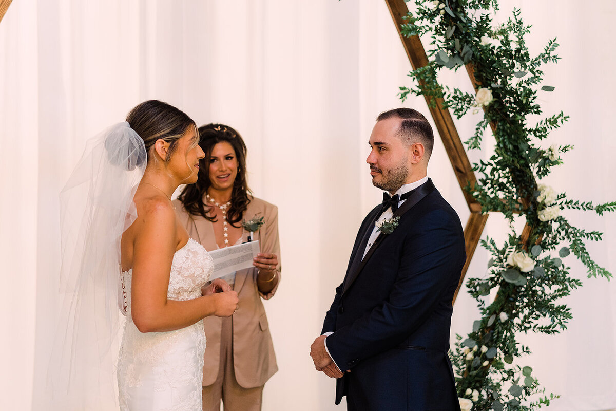 The bride, holding a sheet of vows, gazes at the groom with emotion, as they stand beneath a greenery-adorned arch at their wedding ceremony.