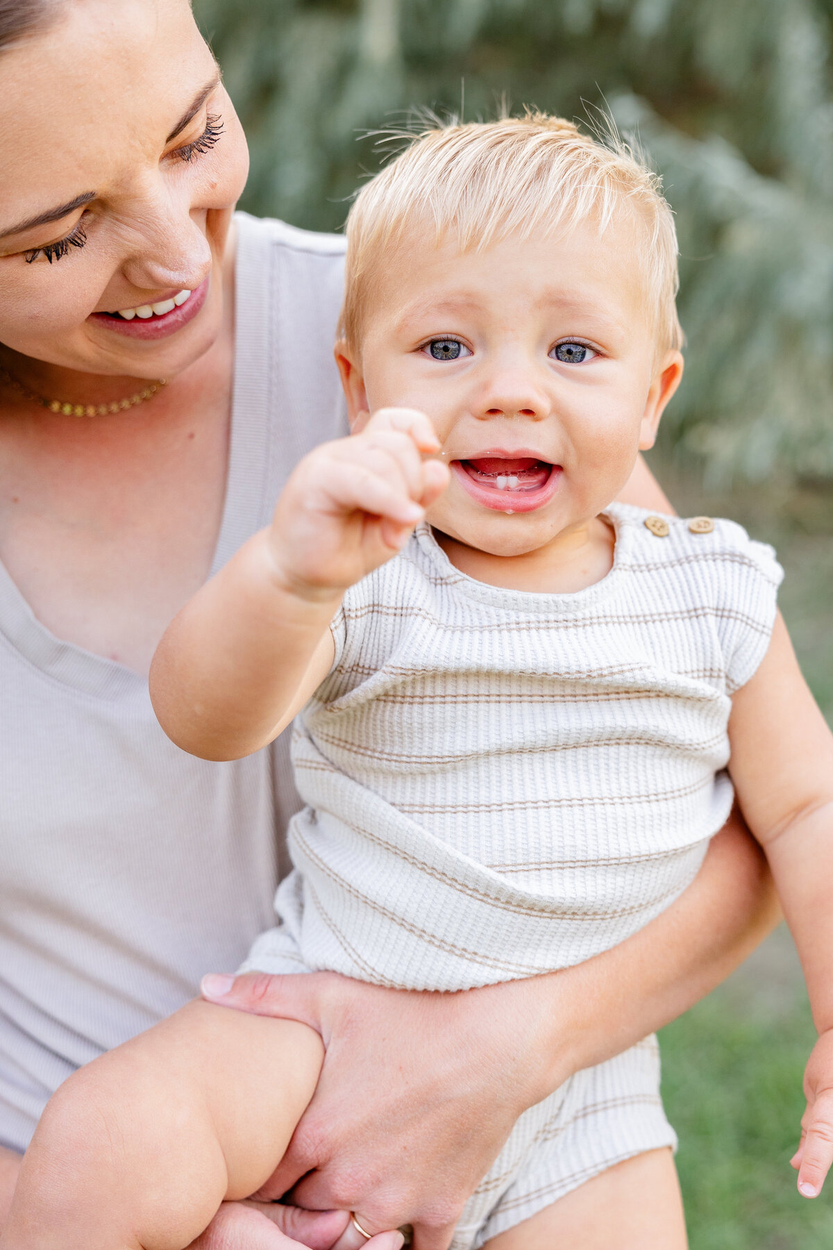 A blonde baby boy giggles while being held by his smiling mother in a peaceful outdoor setting.